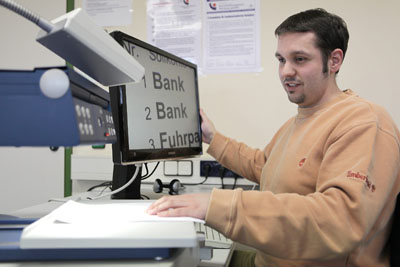 Sehbehinderter junger Mann an einem PC-Arbeitsplatz mit Großbildschirm und Lesegerät