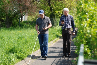 Blinder junger Mann mit Langstock und einem iPhone in der Hand, begleitet von einem Mobilitätstrainer, geht durch einen Grünanlage
