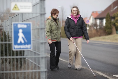 Blinde junge Frau mit Langstock, begleitet von einer Mobilitätstrainerin